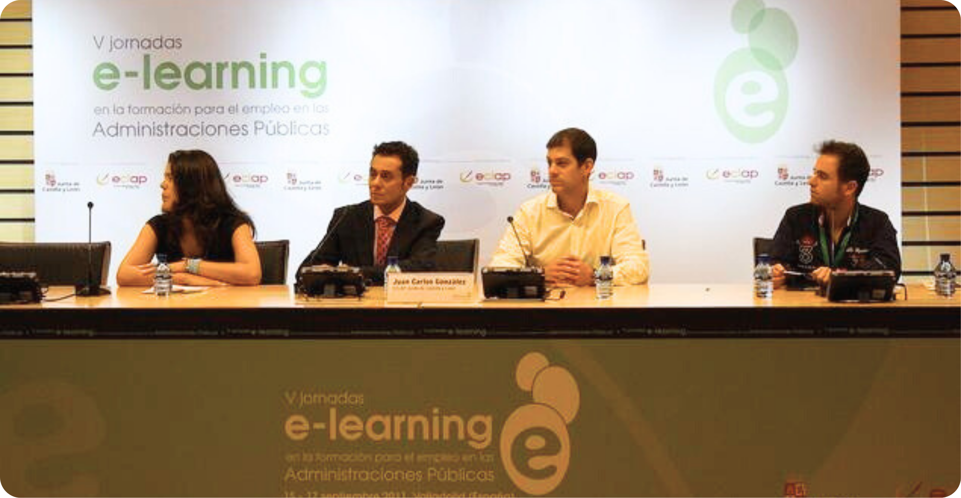 Das Bild zeigt eine Podiumsdiskussion mit vier Personen, die an einem Tisch sitzen. Im Hintergrund befindet sich ein großes Banner mit der Aufschrift "V Jornadas e-learning en la formación para el empleo en las Administraciones Públicas". Auf dem Tisch vor den Personen stehen Mikrofone und Wasserflaschen. Eine der Personen hat ein Namensschild vor sich, auf dem "Juan Carlos González" steht. Das Bild scheint von einer Konferenz oder einem Seminar zu stammen, das sich mit dem Thema E-Learning und dessen Anwendung in öffentlichen Verwaltungen beschäftigt.